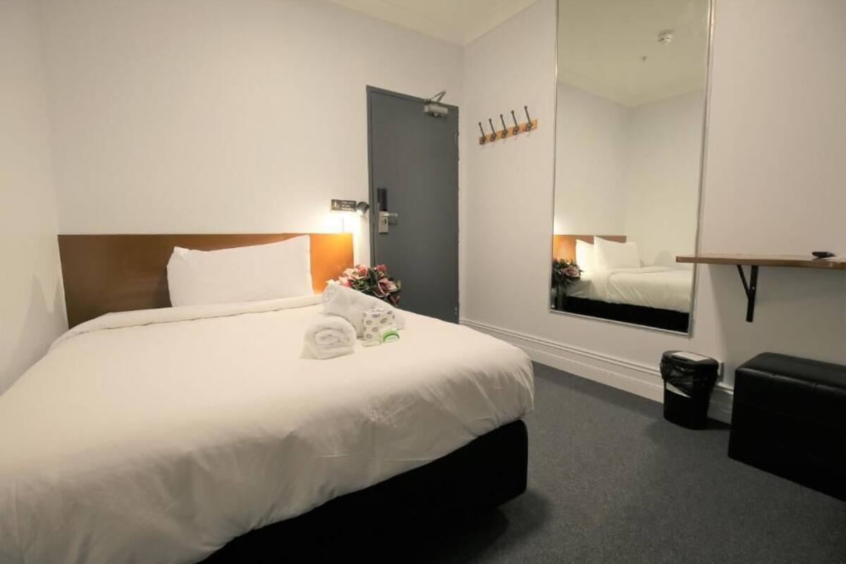 A hotel room with a bed, a mirror, a door, a coat rack, and a trash bin.