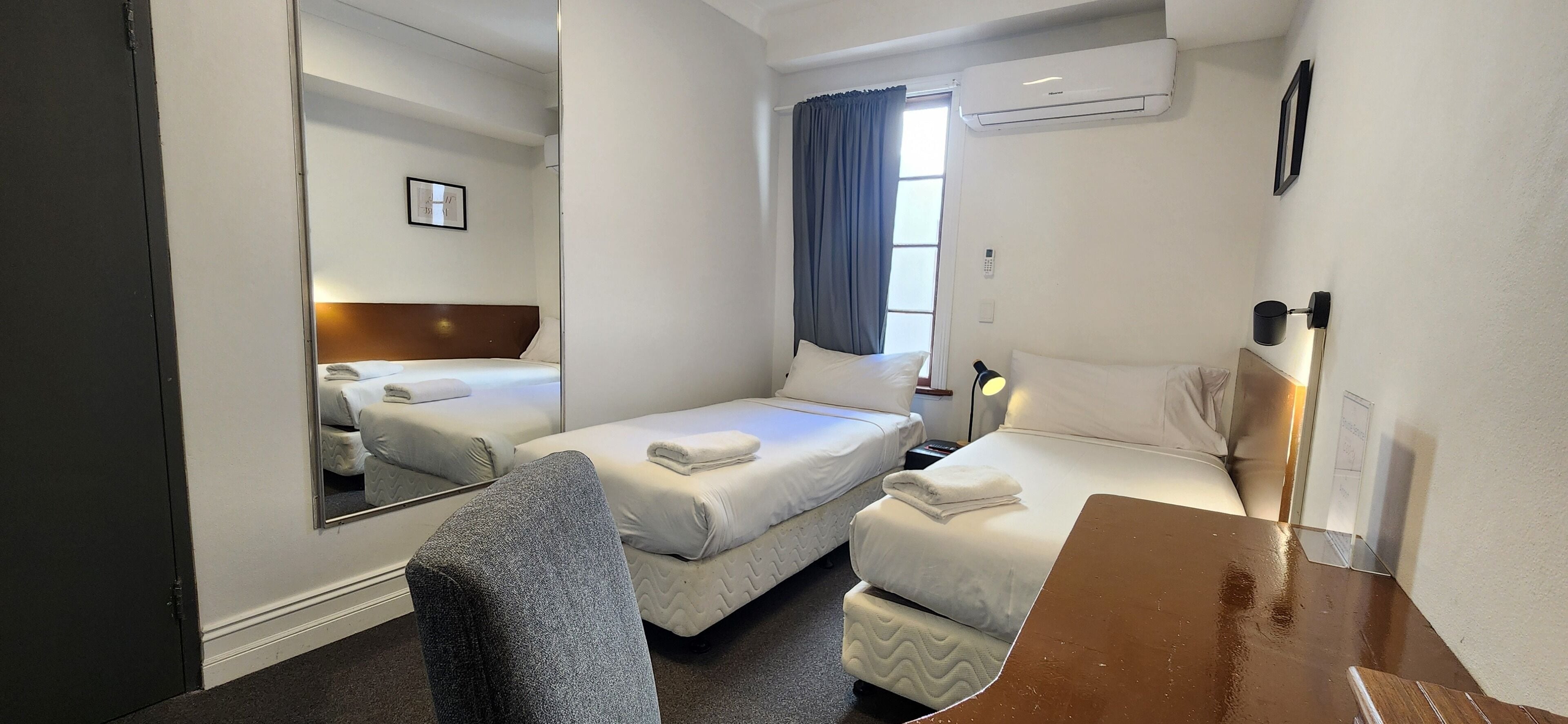 A hotel room with two single beds, a desk, a chair, a mirror, an air conditioning unit, and a window with curtains.