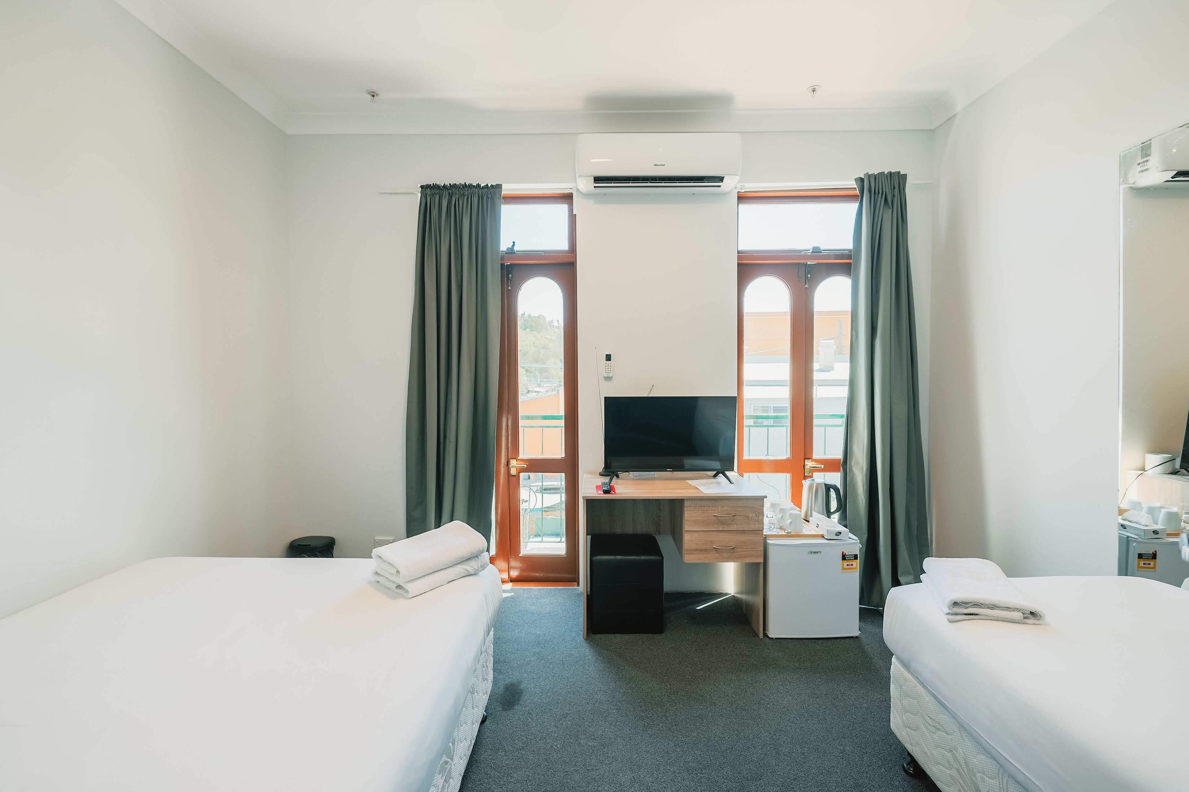 A hotel room with two beds, a desk with a computer, a TV, and a small kitchenette.