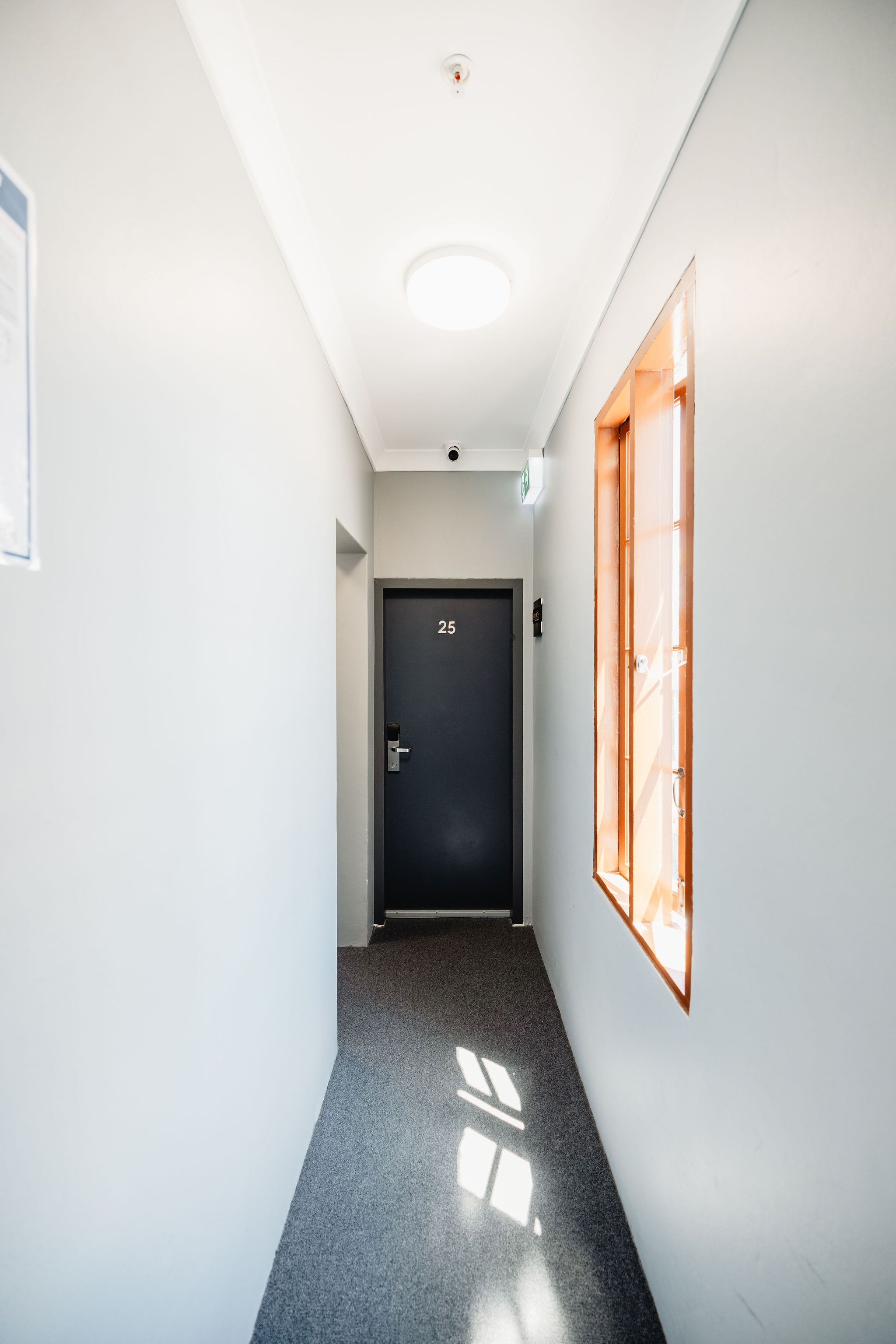 Un couloir étroit avec une porte sombre portant l’inscription « 25 » et une fenêtre aux cadres en bois, située sur le côté droit.