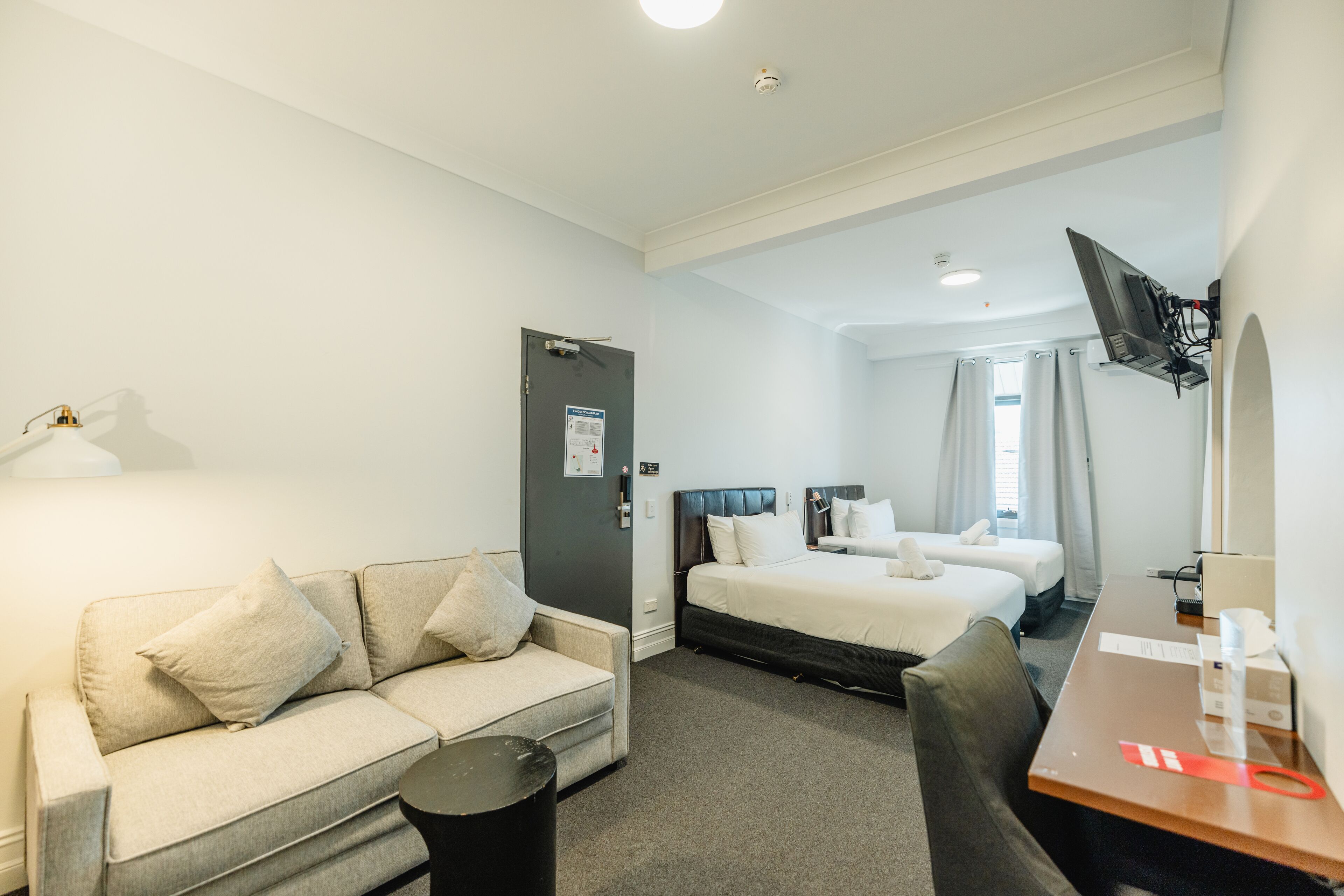 Une chambre d’hôtel avec un canapé, deux lits simples, un bureau, une télévision et une porte avec une pancarte.