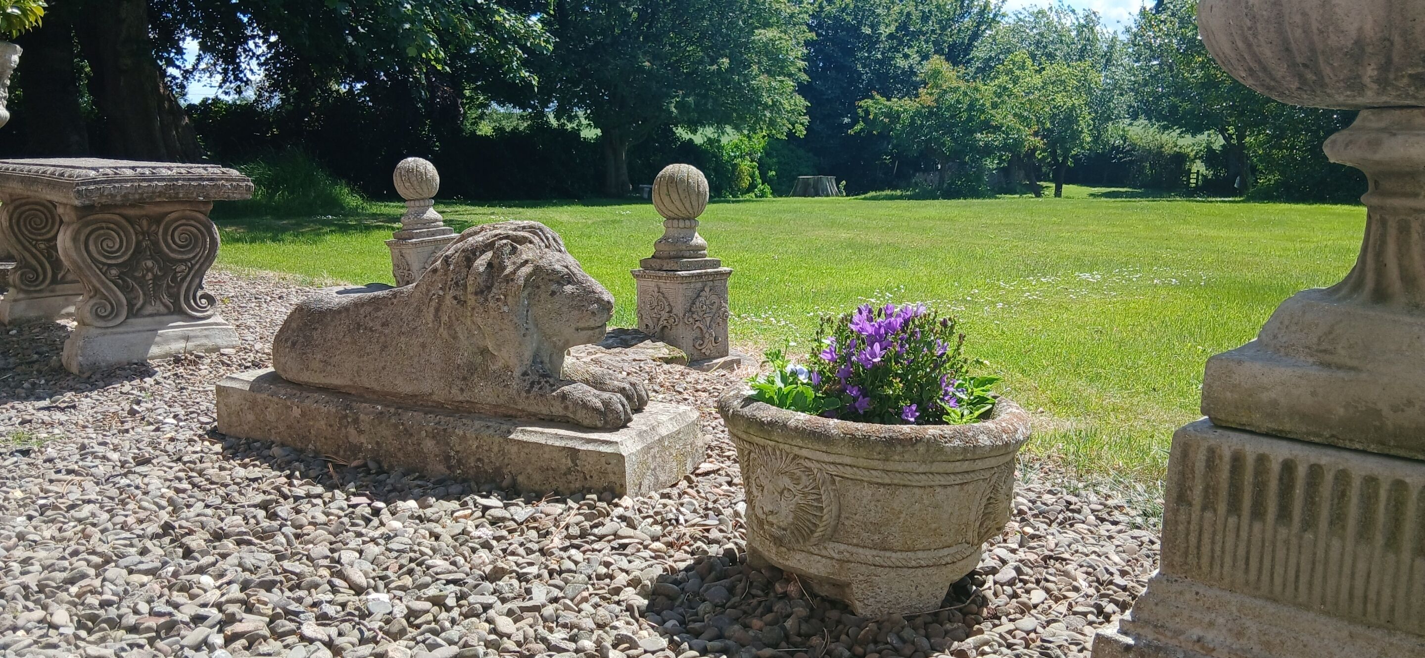 Un jardín con esculturas de piedra, incluyendo un león y una esfinge, además de una planta en maceta con flores moradas.