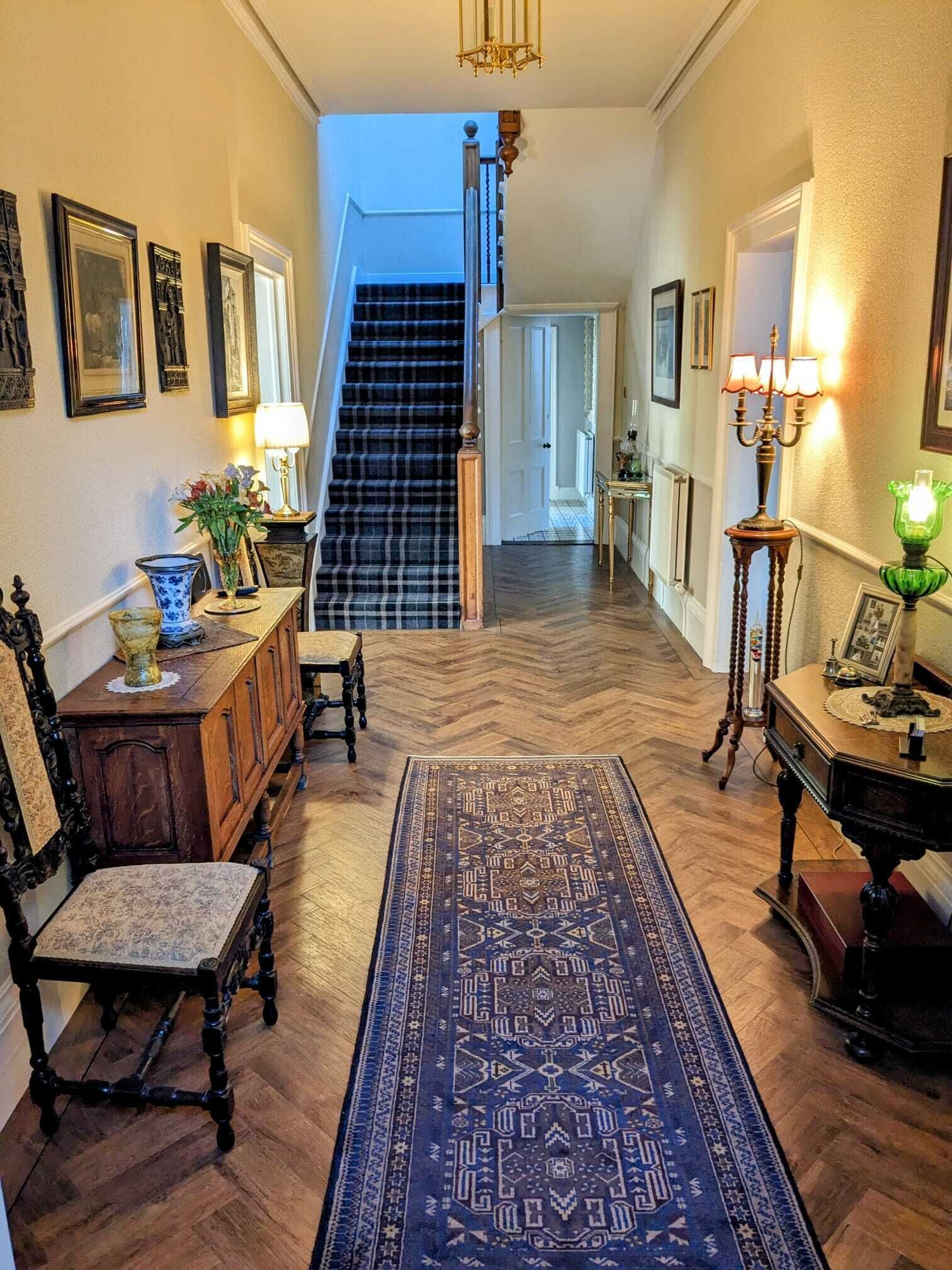 En hall med ett fiskbensmönstrat golv, en blå, mönstrad matta, en trästol, ett bord med en lampa och en trappa som leder upp till en våning ovanför.