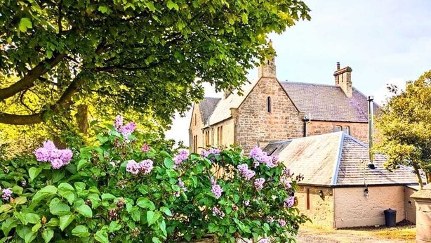 A stone building with a traditional roof, surrounded by lush greenery and flowering plants. - The Gables (Berwick-upon-Tweed)
