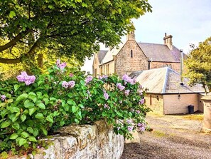 A stone building with a traditional roof, surrounded by lush greenery and flowering plants. - The Gables (Berwick-upon-Tweed)
