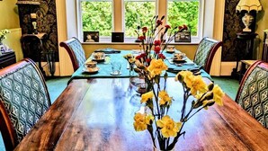 A dining room with a wooden table set for four, flanked by patterned chairs, with a window offering a view of greenery. - The Gables (Berwick-upon-Tweed)