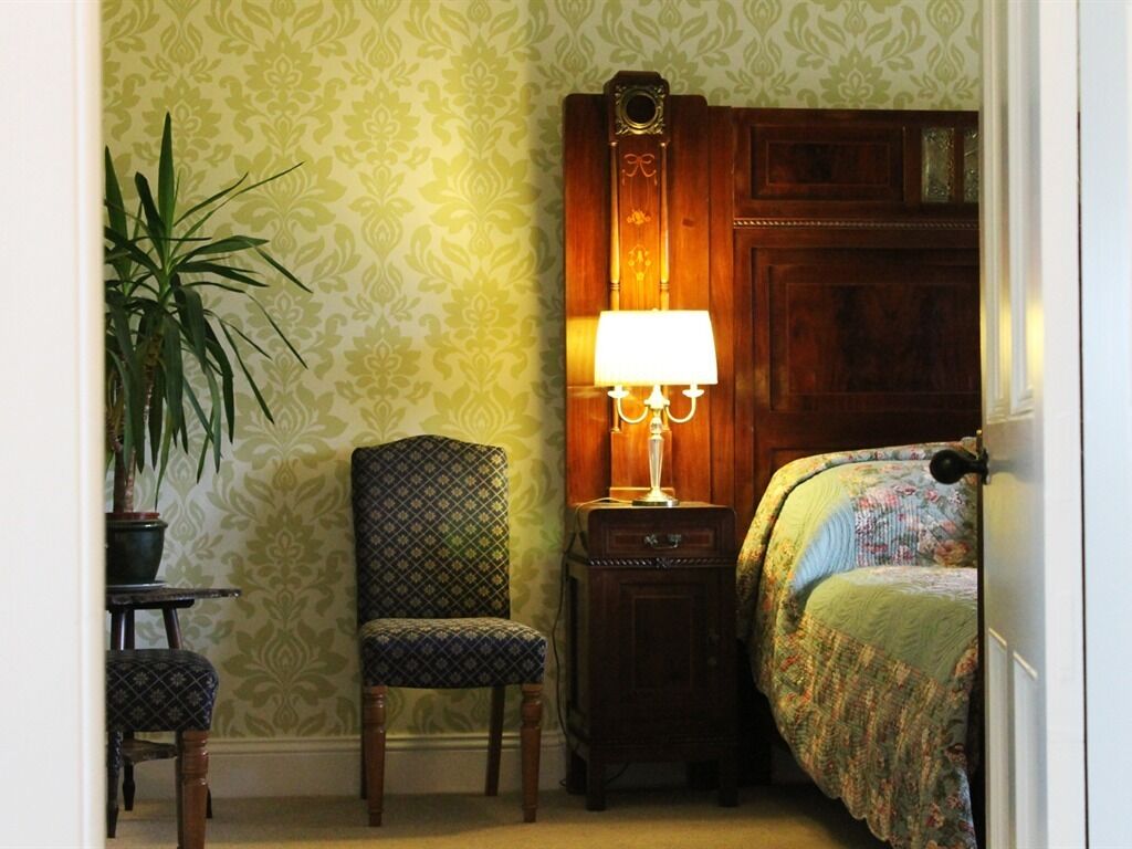 A corner room with a patterned wallpaper, a wooden nightstand with a lamp, a patterned chair, and a bed with a floral bedspread.