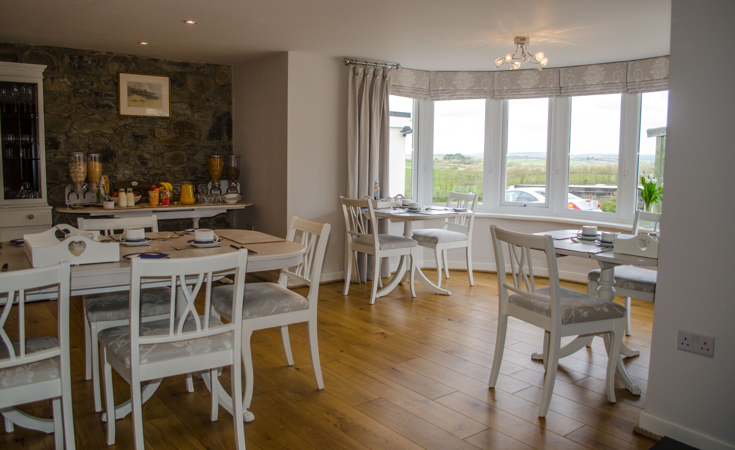 Un coin repas avec des chaises blanches et une table, un mur en pierre orné d’un tableau, et de grandes fenêtres offrant une vue sur la campagne.