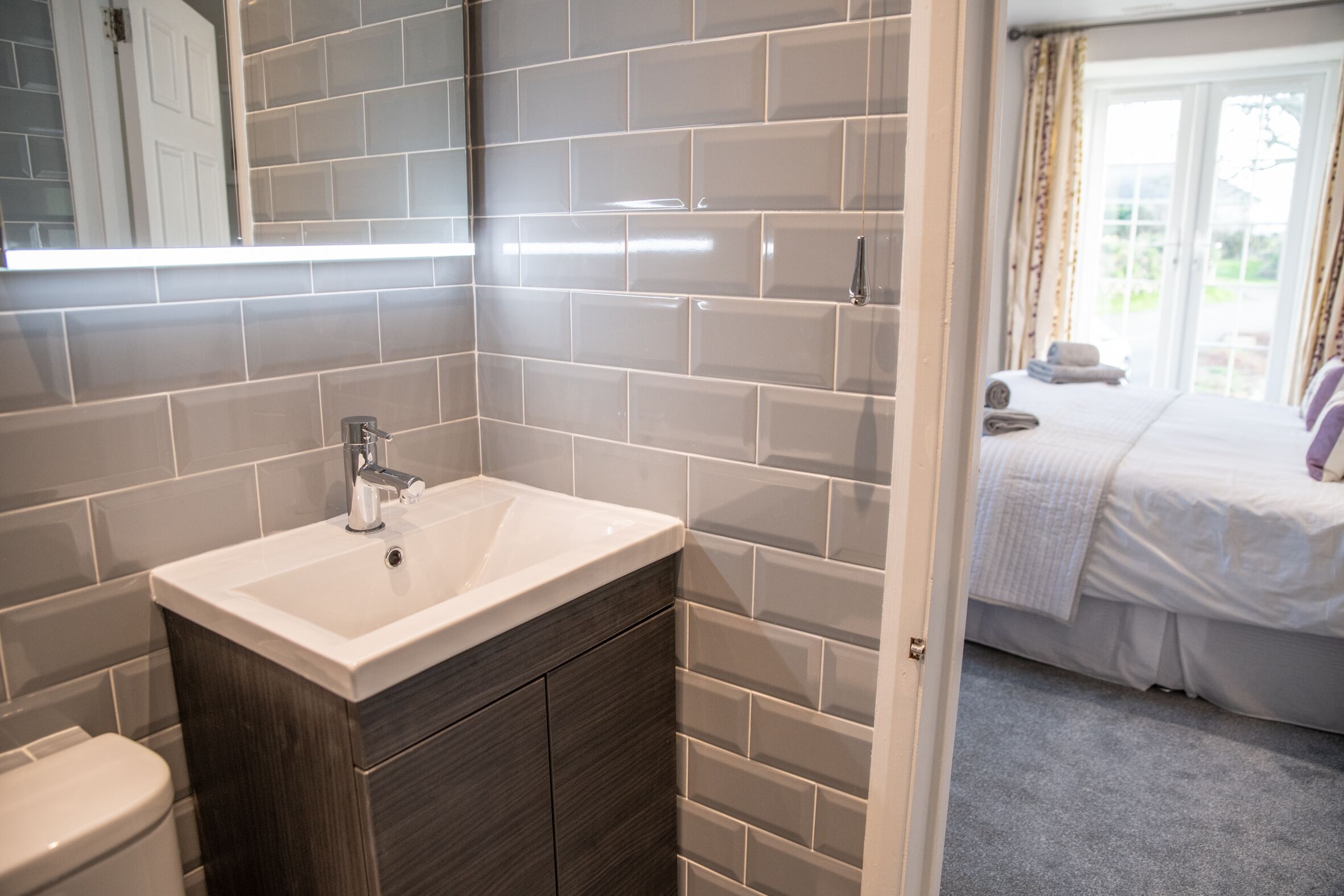 Un baño con lavamanos blanco, armario de madera oscura y paredes con azulejos grises.