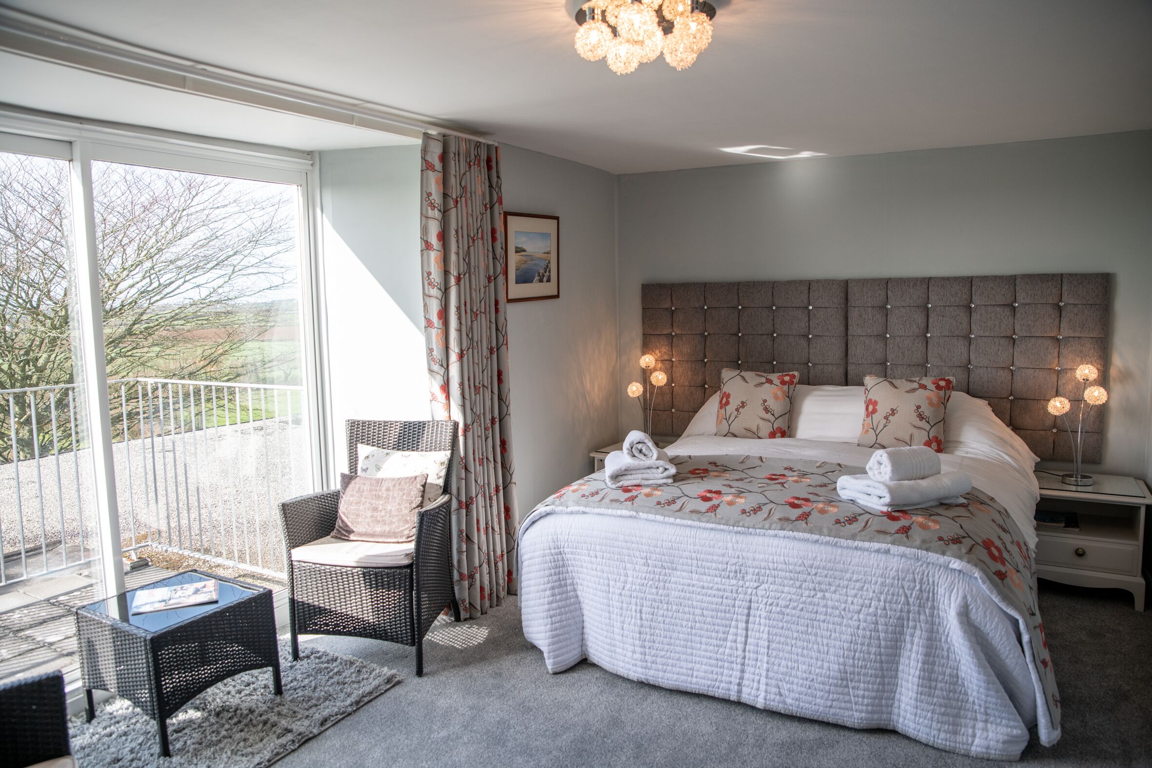 A bedroom with a large bed, a nightstand, a chair, and a view of the outdoors.