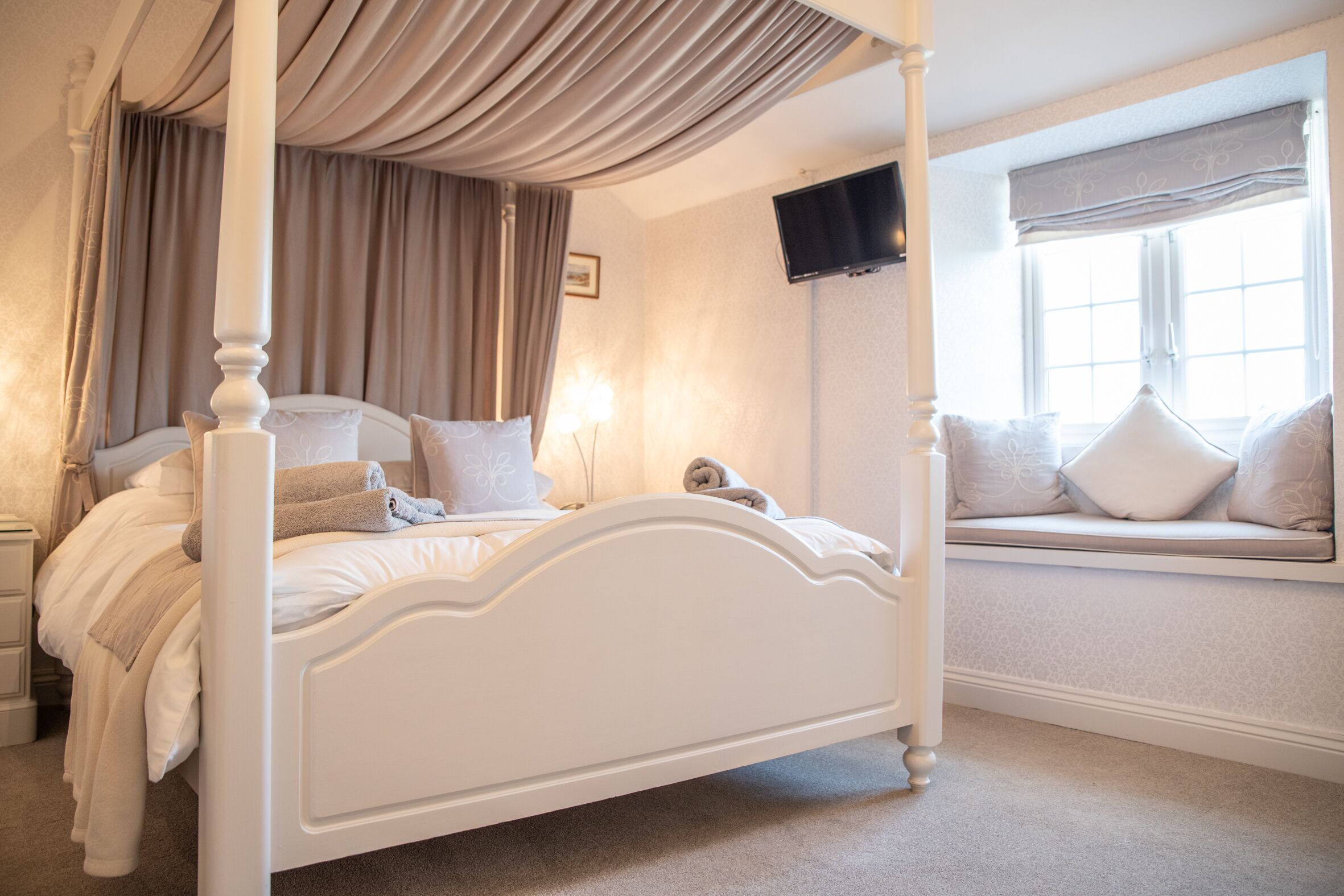 A four-poster bed with a canopy, a bedside table, a window seat with cushions, and a flat-screen TV mounted on the wall.