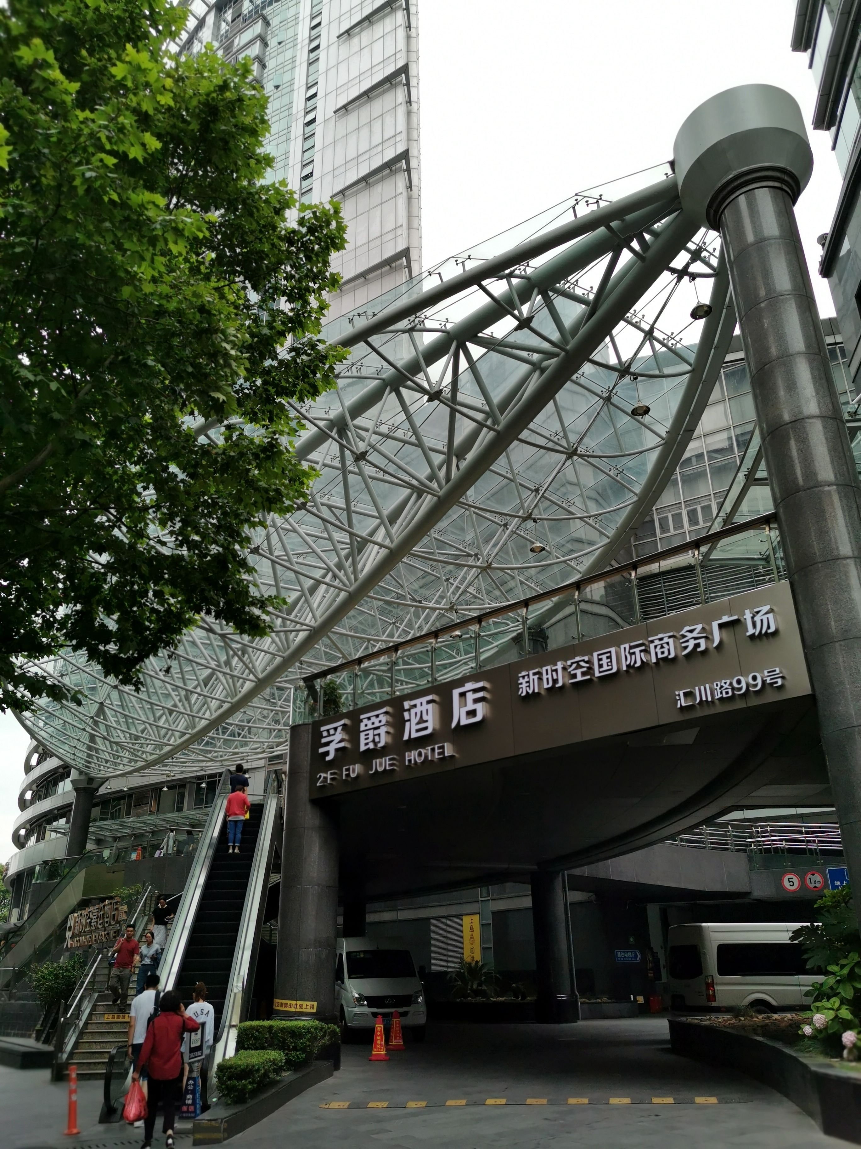 A hotel entrance with a modern glass and steel structure, an escalator, and a sign indicating the hotel name and address.