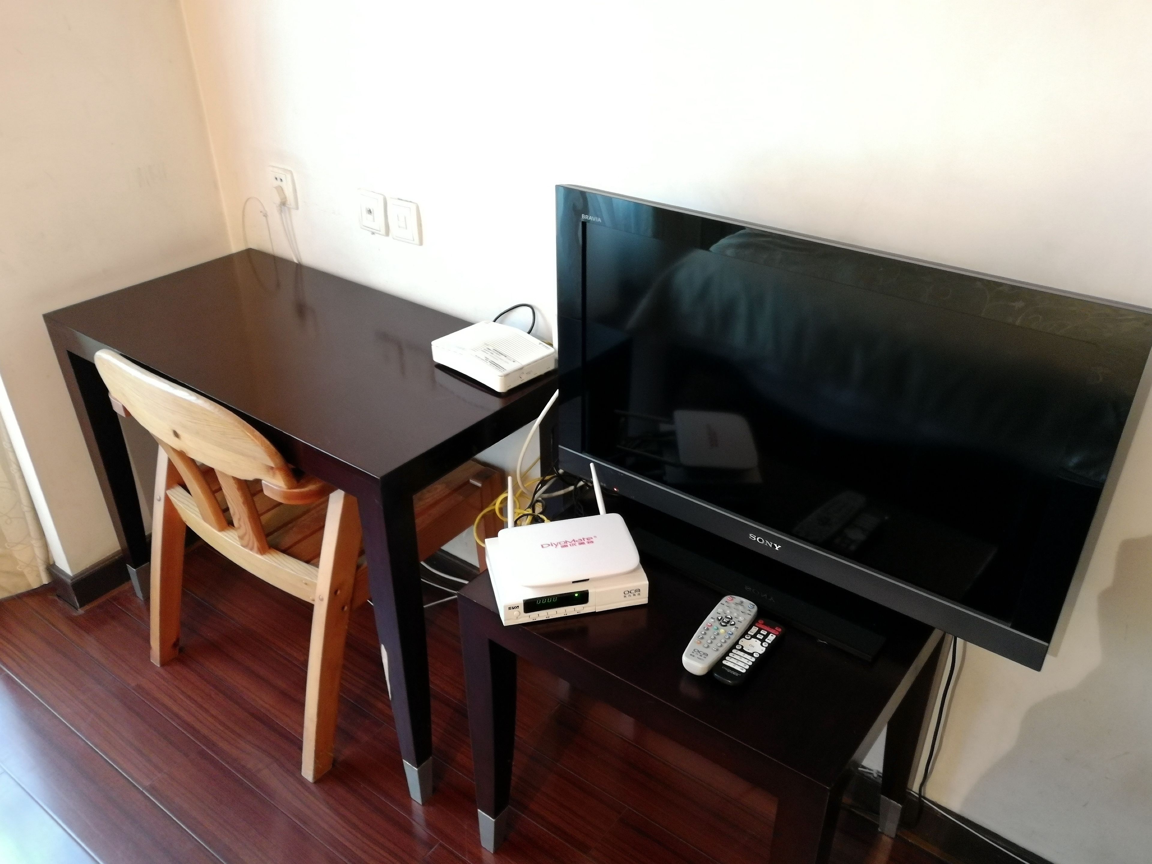 A dark wooden desk with a flat-screen TV, a wooden chair, a white power strip, and two remote controls.