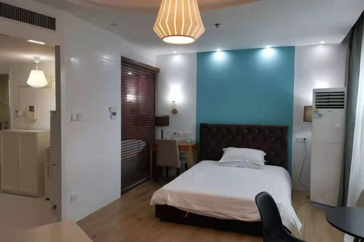 A modern bedroom with a bed, a desk, and a chair. There is a window with blinds, a ceiling light, and an air conditioning unit.