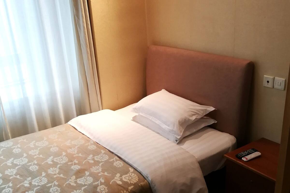 Ein Einzelbett mit Kopfteil, ein Nachttisch mit Fernbedienung und ein Fenster mit Vorhängen.