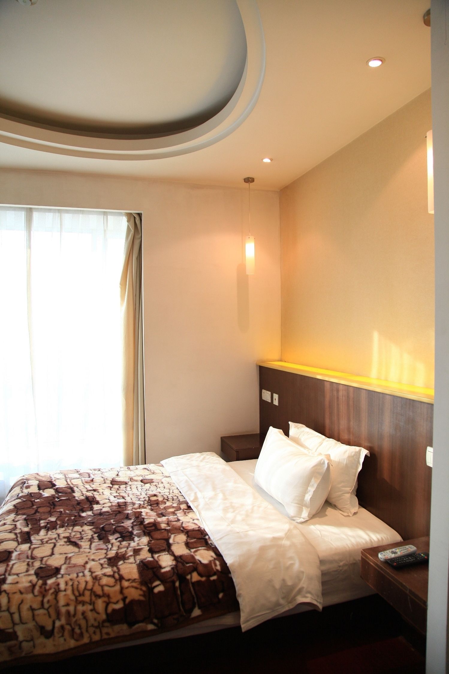 Una habitación de hotel con cama, mesita de noche, control remoto y ventana con cortinas.