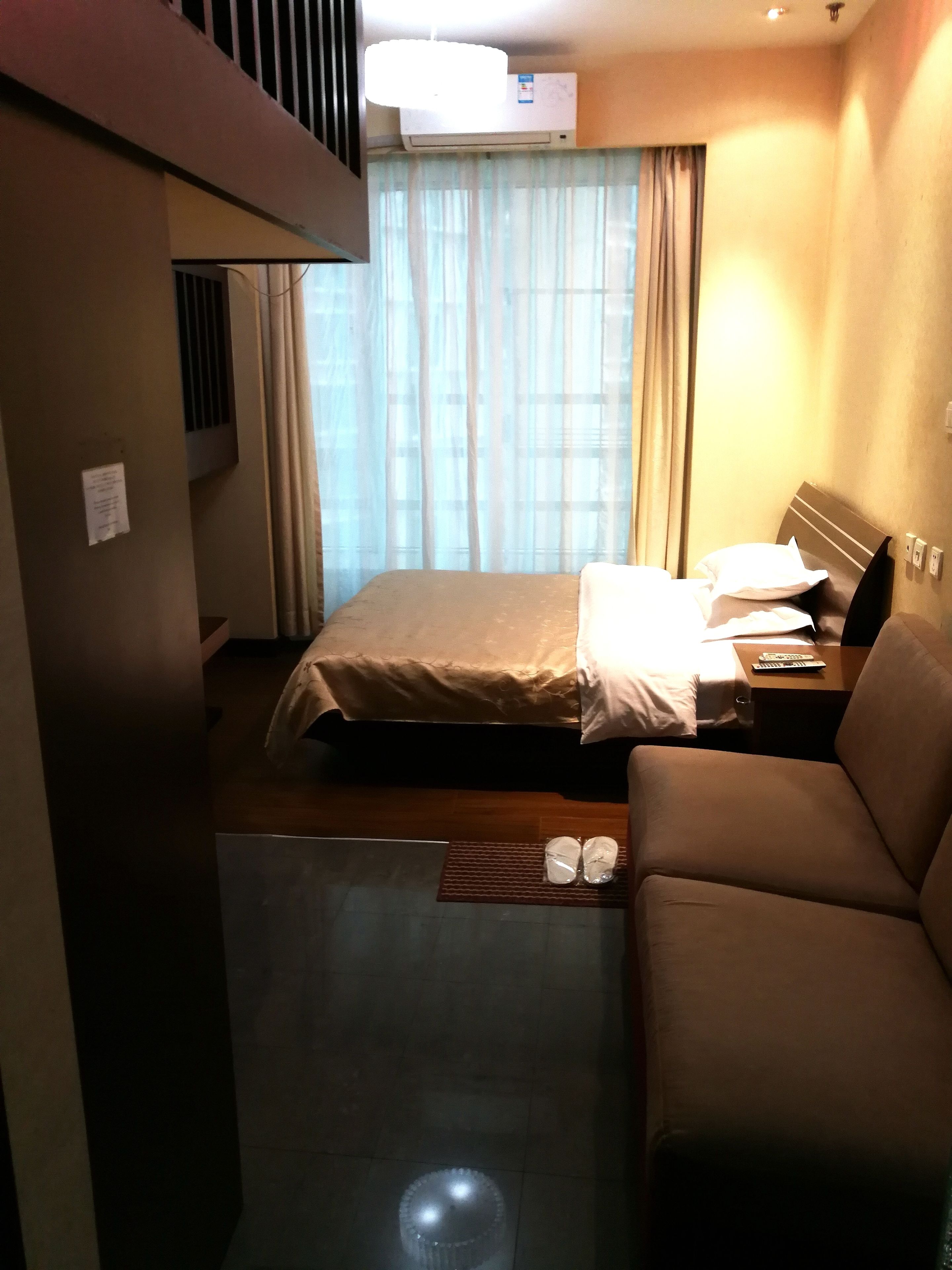 A hotel room with a bed, a sofa, a small table with a remote control, and an air conditioning unit.