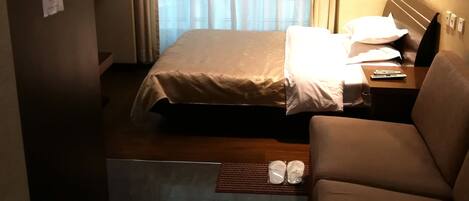 A hotel room with a bed, a sofa, a small table with a remote control, and an air conditioning unit.