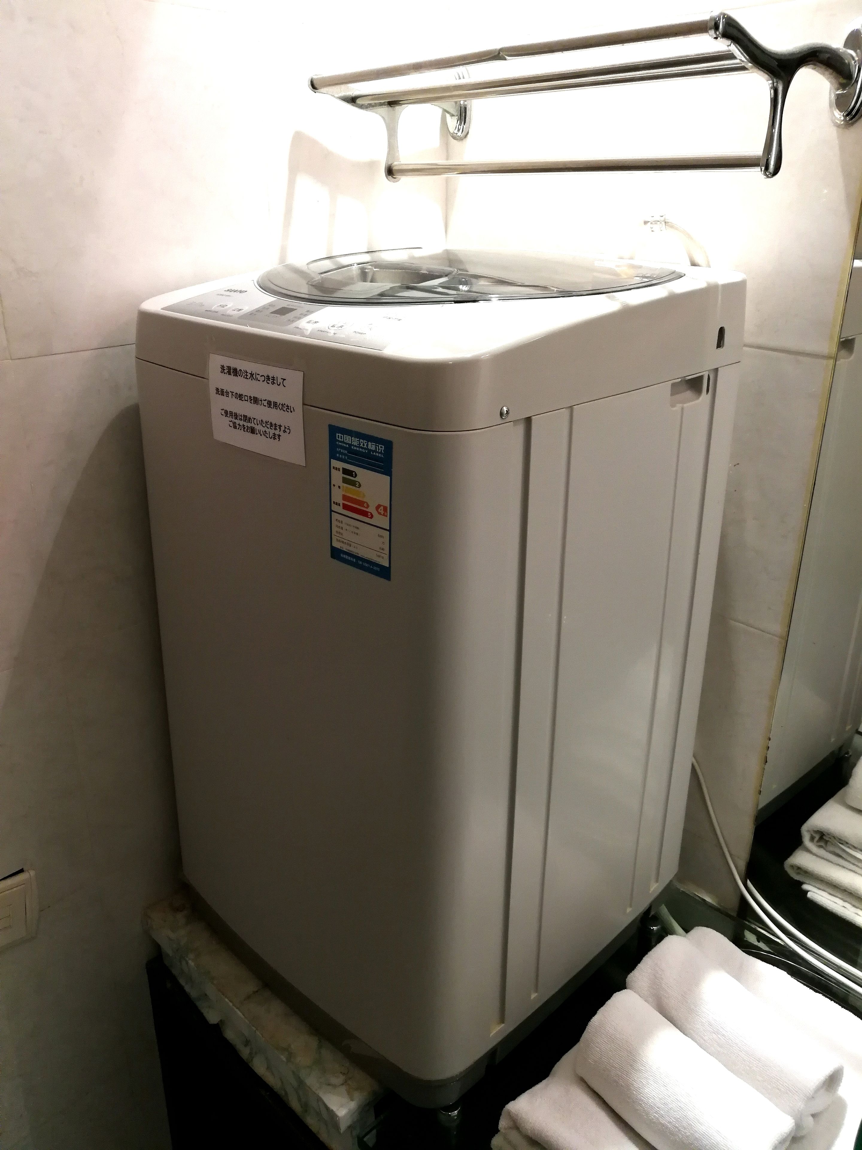 A compact, white, front-loading washing machine in a bathroom with a metal drying rack above it and a towel rack to the right.