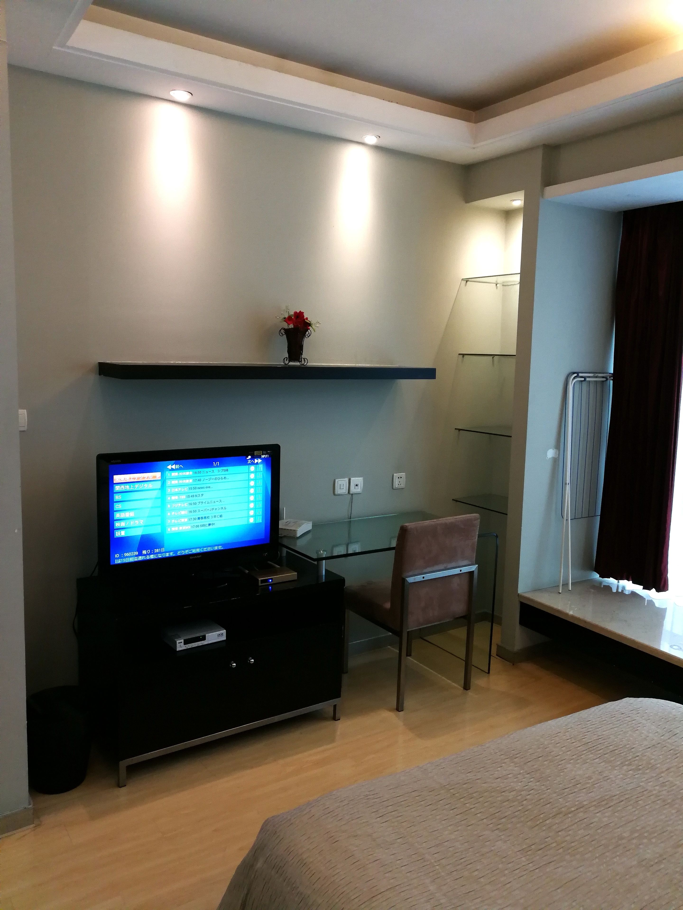 A hotel room with a bed, a desk with a computer, a chair, a TV, and a small shelf with a flower.