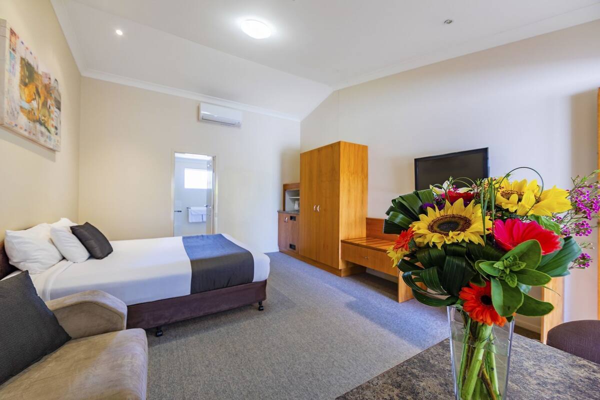A hotel room with a bed, a sofa, a wooden wardrobe, a desk, a TV, and a vase of flowers.