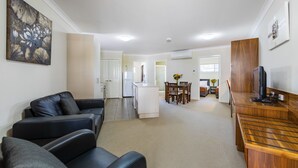 Una sala moderna con un sofá de cuero negro, una mesa de centro de madera y un televisor. Se ve una cocina al fondo, con armarios blancos, y un comedor con una mesa y sillas de madera.