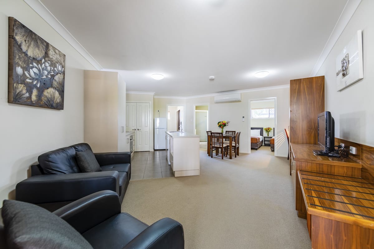 A modern living room with a black leather sofa, a wooden coffee table, and a TV. There is a kitchen in the background with white cabinets and a dining area with a wooden table and chairs.