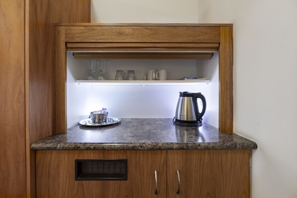 A kitchen area with a granite countertop, a coffee maker, a kettle, and a shelf with glassware.
