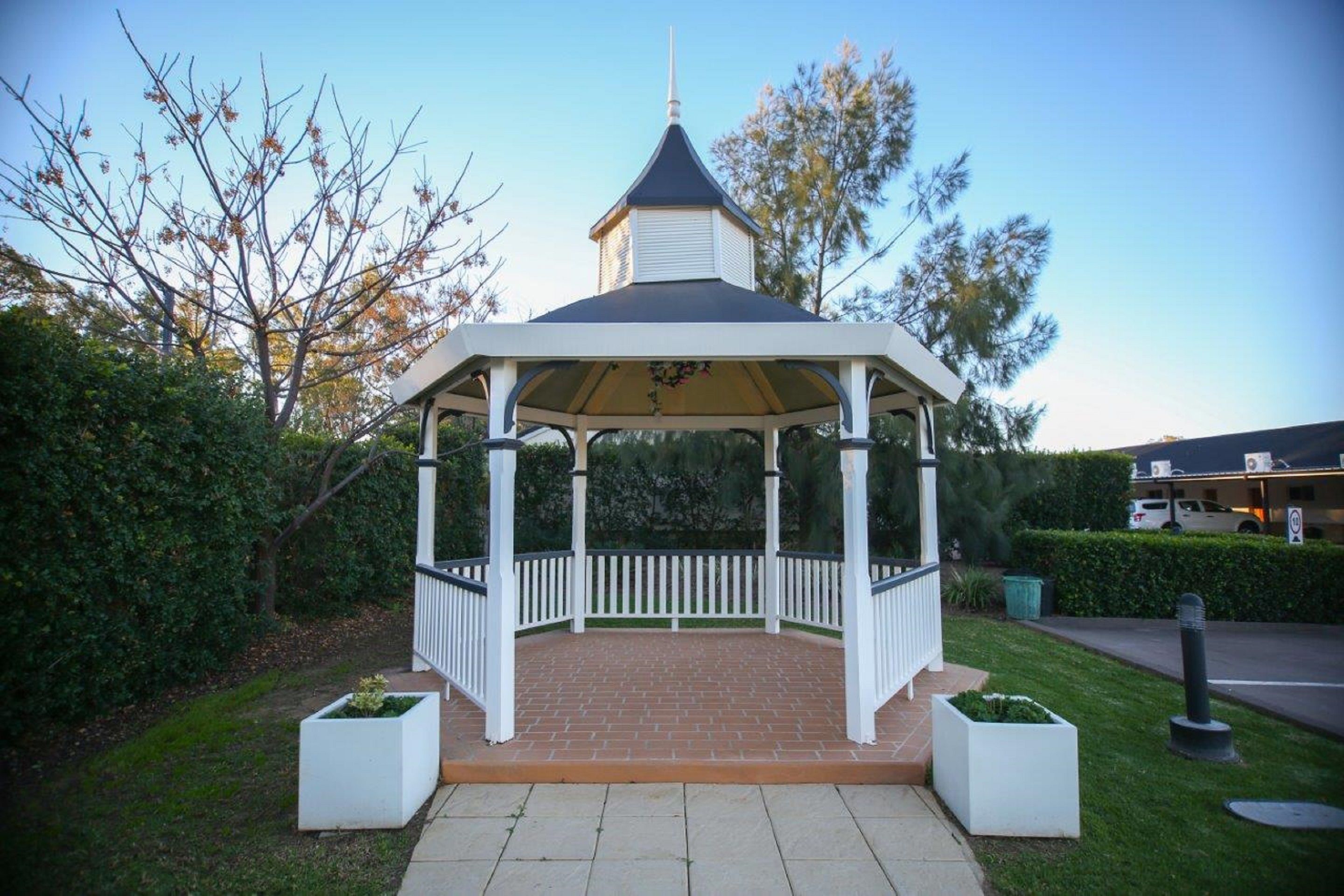 A gazebo with a white roof and a pointed top, surrounded by a white picket fence, set on a brick patio with planters on either side.