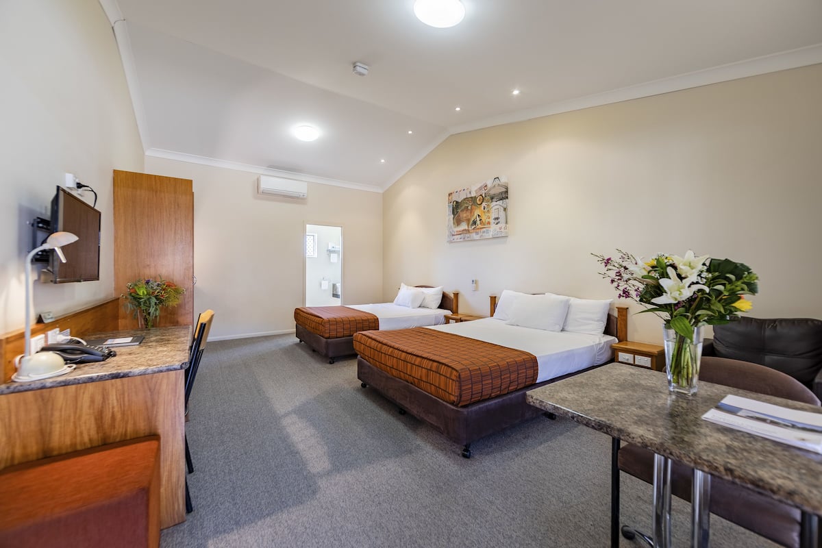 A hotel room with two beds, a desk with a chair, a small table, a vase with flowers, and a painting on the wall.