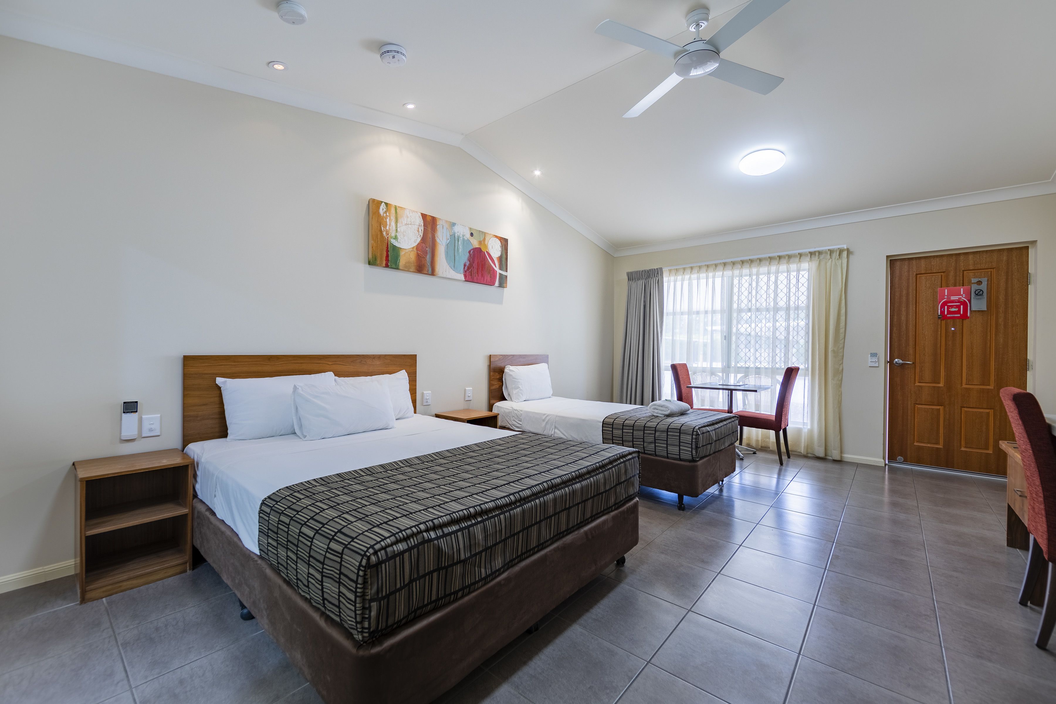 A hotel room with two beds, a ceiling fan, a painting, a window with curtains, and a door with a sign.