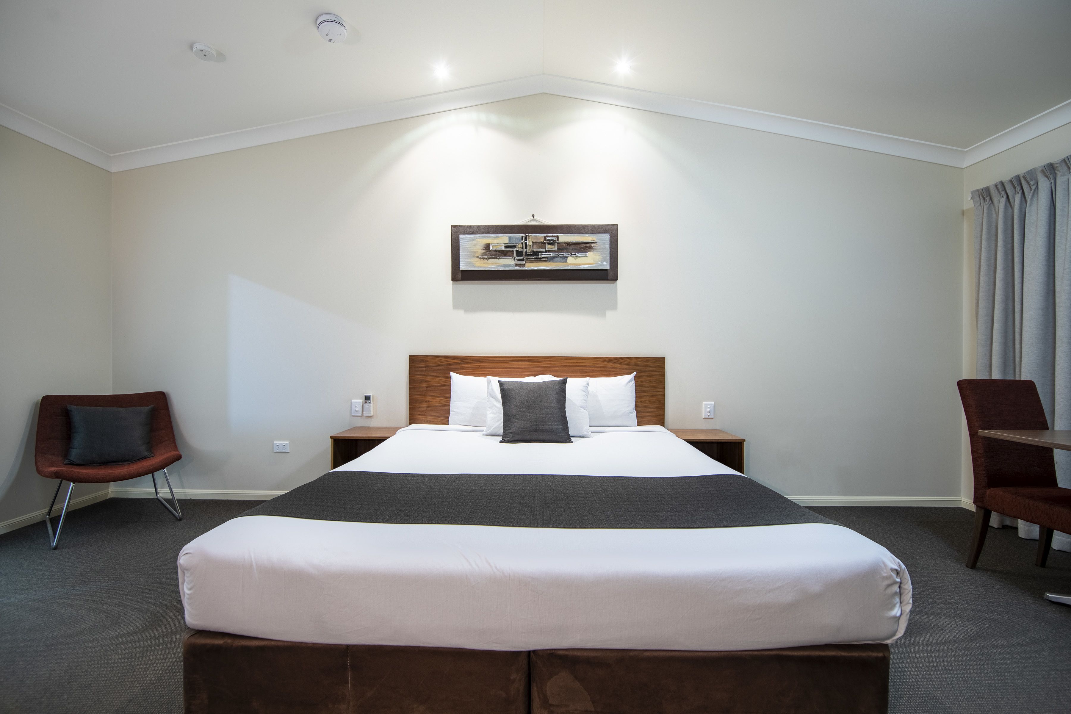 Habitación de hotel con una cama grande, cabecera, mesita de noche, una silla y un cuadro en la pared.