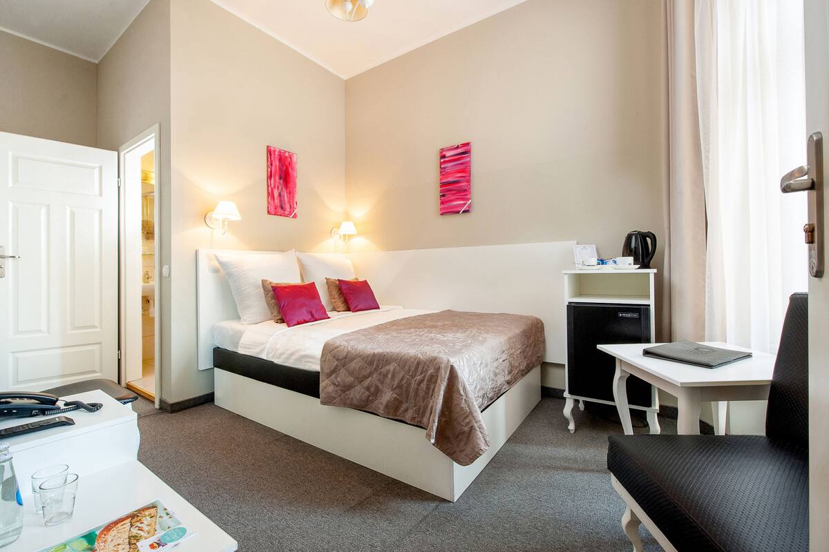 A hotel room with a bed, bedside table, lamp, and a small desk with a chair.