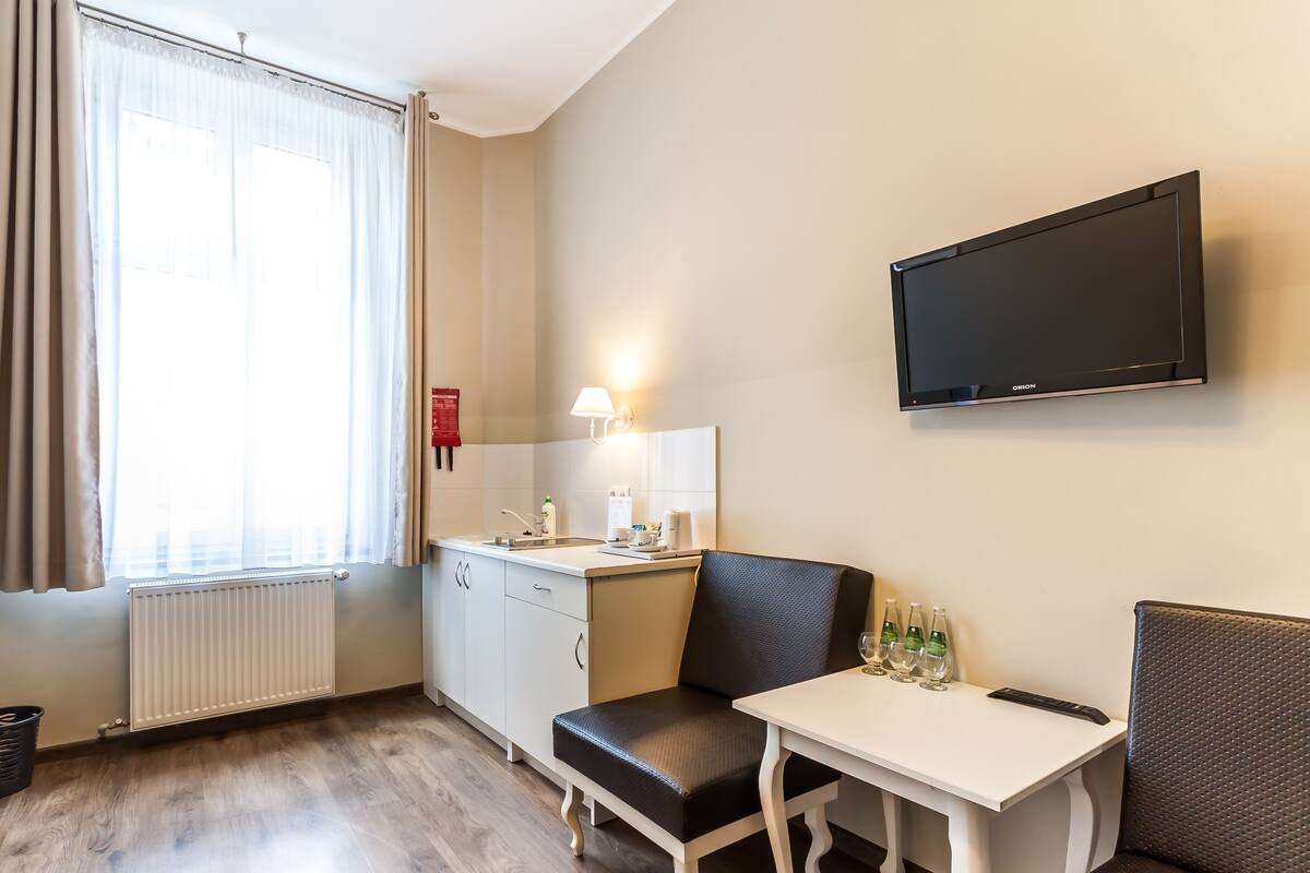 A hotel room with a kitchenette, a dining area with a table and chairs, a flat-screen TV mounted on the wall, and a window with curtains.