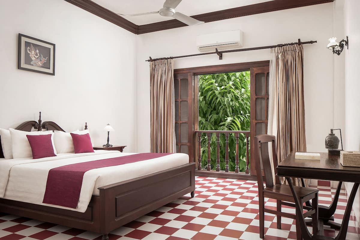Quarto com cama, mesa de cabeceira, uma mesa e uma cadeira. Há uma janela com cortinas e vista para o verde lá fora.