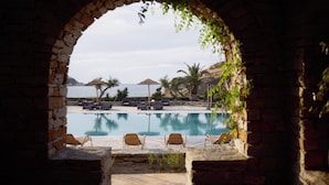 Une vue sur la piscine, avec des chaises longues et des parasols, une vue dégagée sur la mer et une arche en pierre qui encadre le paysage.