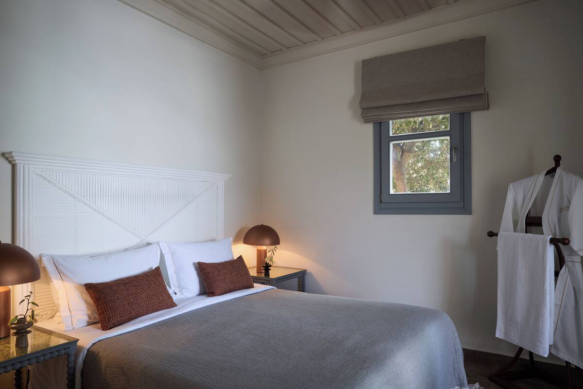 Un dormitorio con cama, lámparas de noche, ventana con vistas y perchero para bata.
