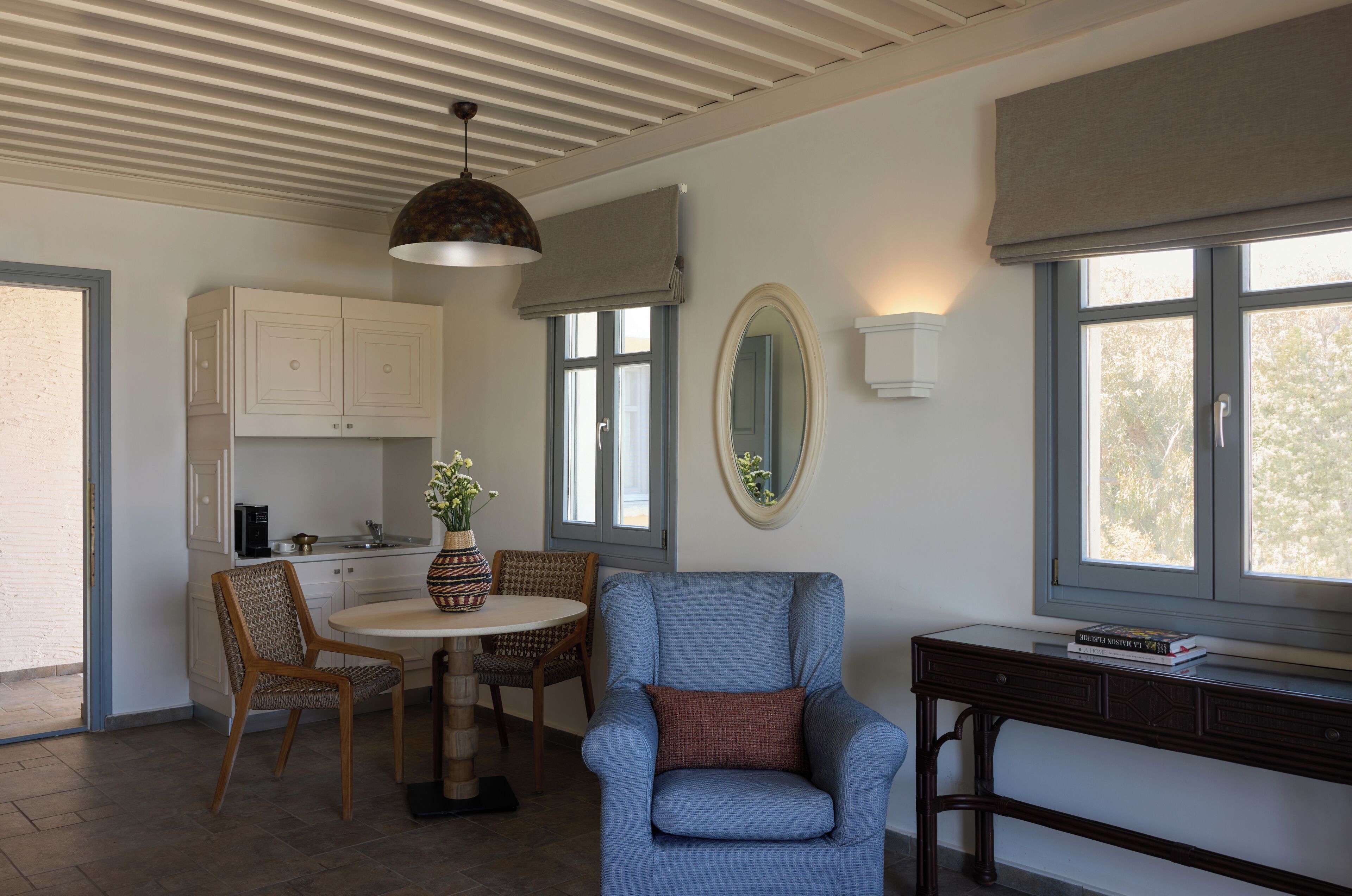 Une pièce comprenant une table à manger, des chaises, un fauteuil bleu, un petit bureau, un miroir et une fenêtre donnant sur un paysage agréable.