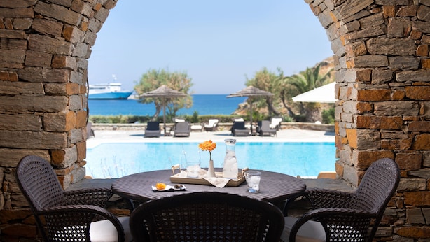 Un arc en pierre surplombe une piscine agrémentée de chaises longues et de parasols, une table avec des boissons, et offre une vue sur la mer.