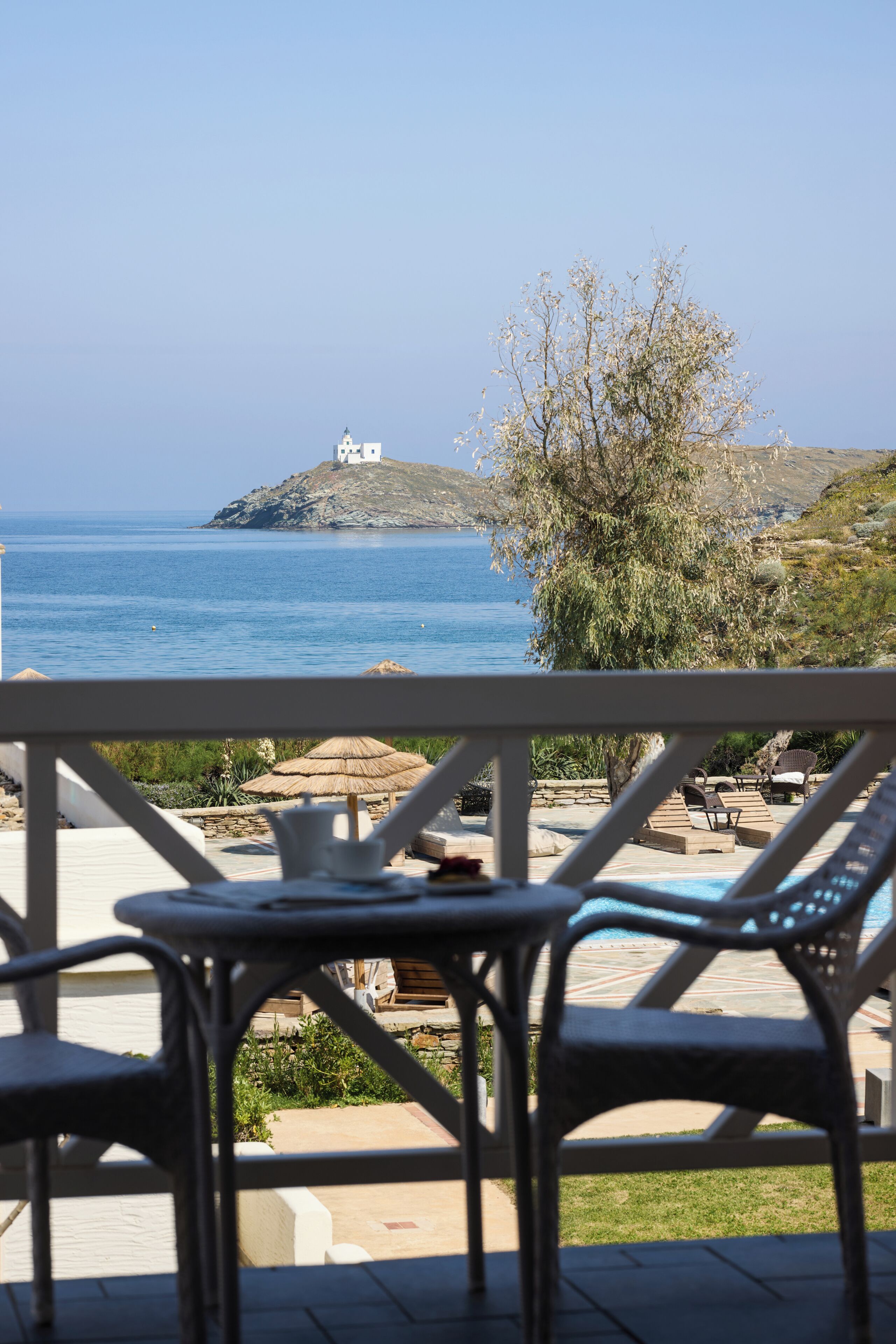 A seaside view with a white building on a hill, a pool area with lounge chairs, and a clear blue sky.