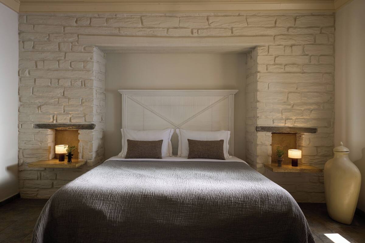 Un dormitorio con pared de piedra, una cama con cabecera, dos mesitas de noche con lámparas y un jarrón grande a la derecha.