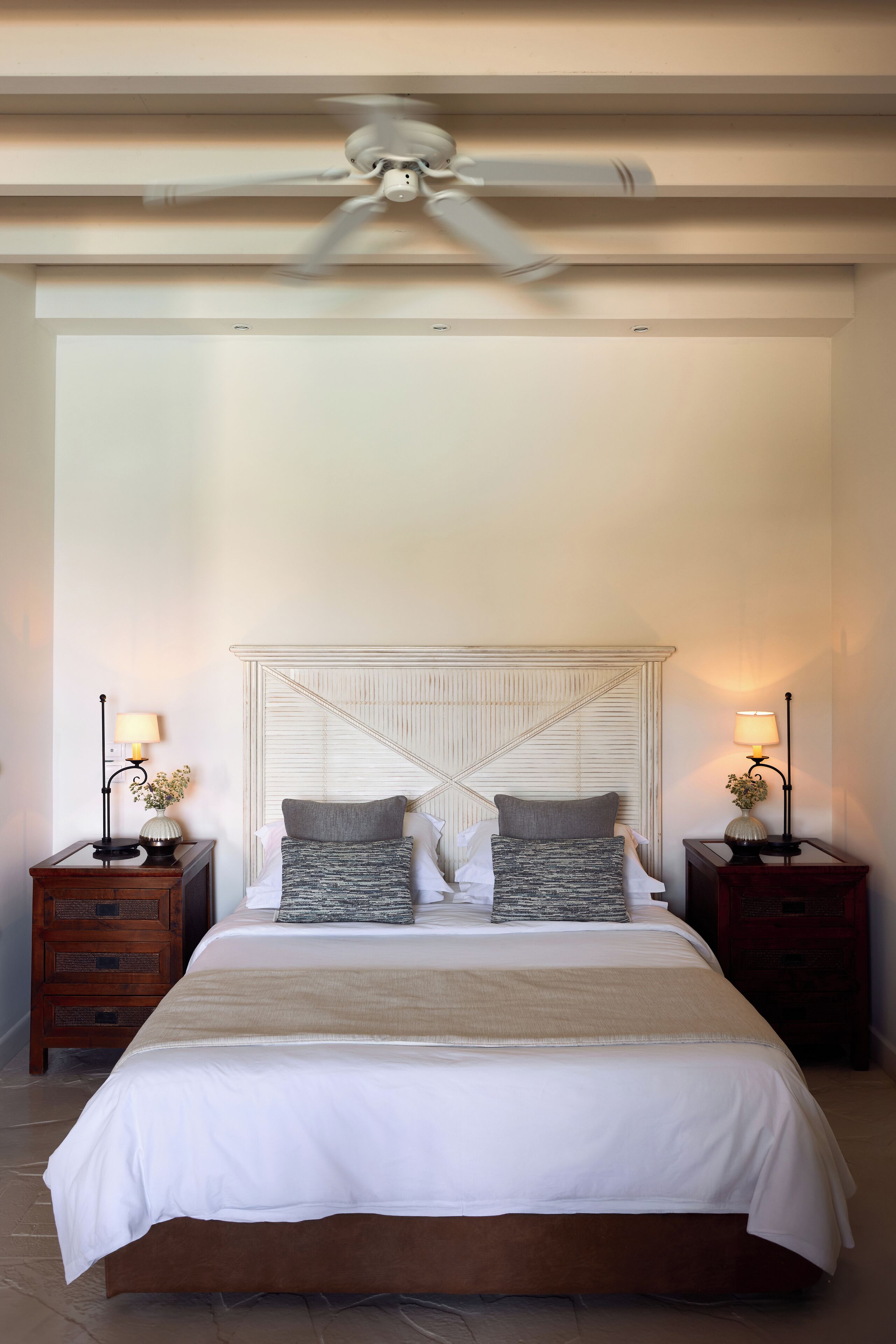 Un dormitorio con una cama grande, dos mesitas de noche con lámparas, un ventilador de techo y un cabecero texturizado.
