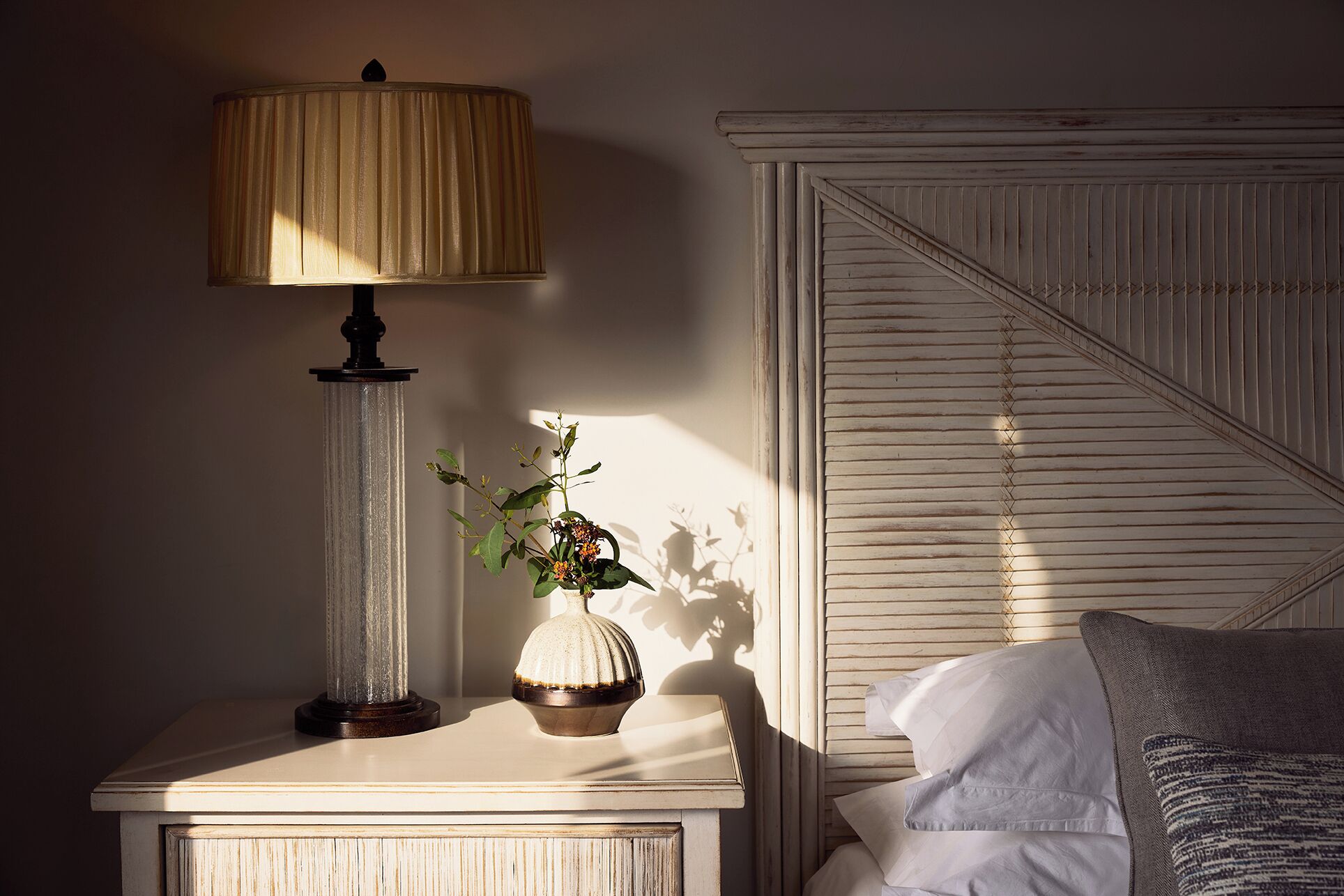 A bedroom with a nightstand, a lamp, a vase with flowers, and a bed with pillows.