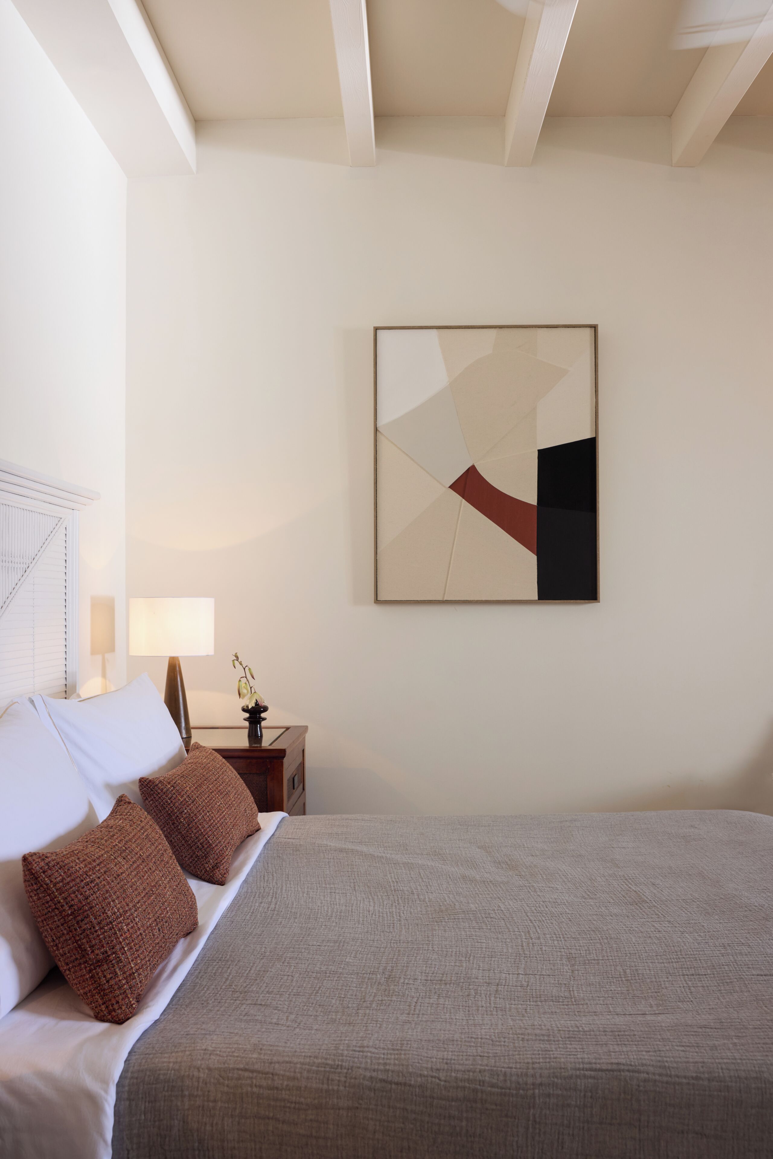 A bedroom with a bed, pillows, a nightstand, a lamp, and a wall-mounted abstract painting.