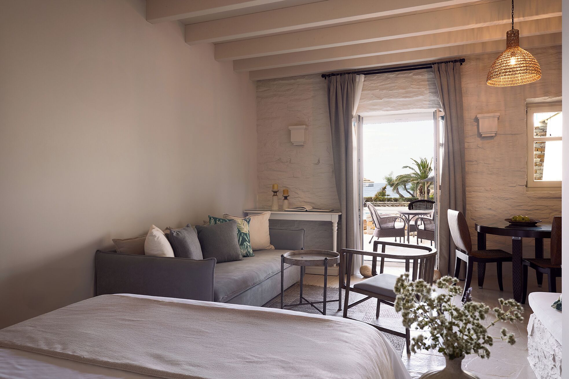 Une chambre d’hôtel moderne comprenant un lit, un coin salon avec une table et des chaises, une petite table ronde et une vue sur la mer par une grande fenêtre.