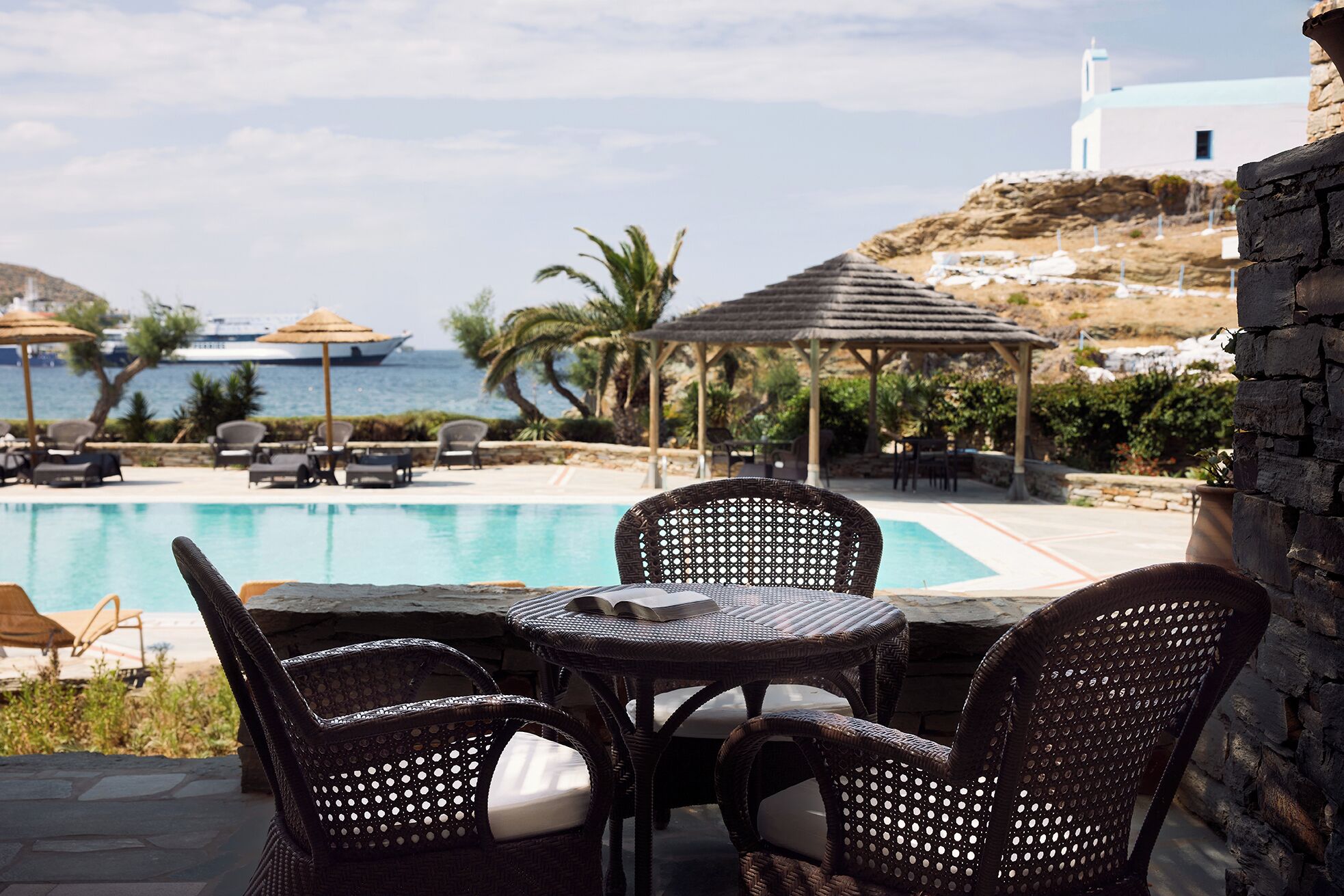 Zona junto a la piscina con sillas de mimbre y una mesa, con vistas a una piscina y al paisaje costero, con un edificio blanco al fondo.