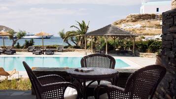 Zona junto a la piscina con sillas de mimbre y una mesa, con vistas a una piscina y al paisaje costero, con un edificio blanco al fondo.
