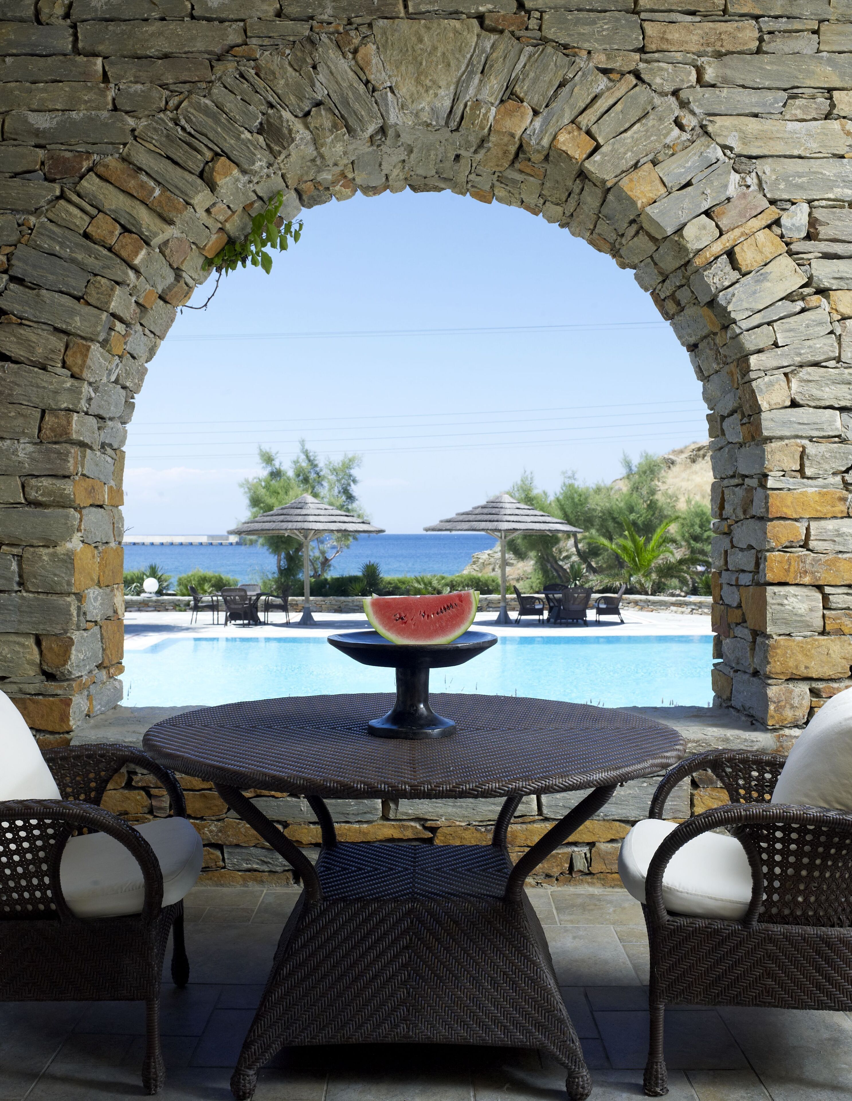 Ein Sitzbereich im Freien mit einem steinernen Bogen, einem Tisch mit einer Wassermelone als Mittelpunkt und einem Pool im Hintergrund.