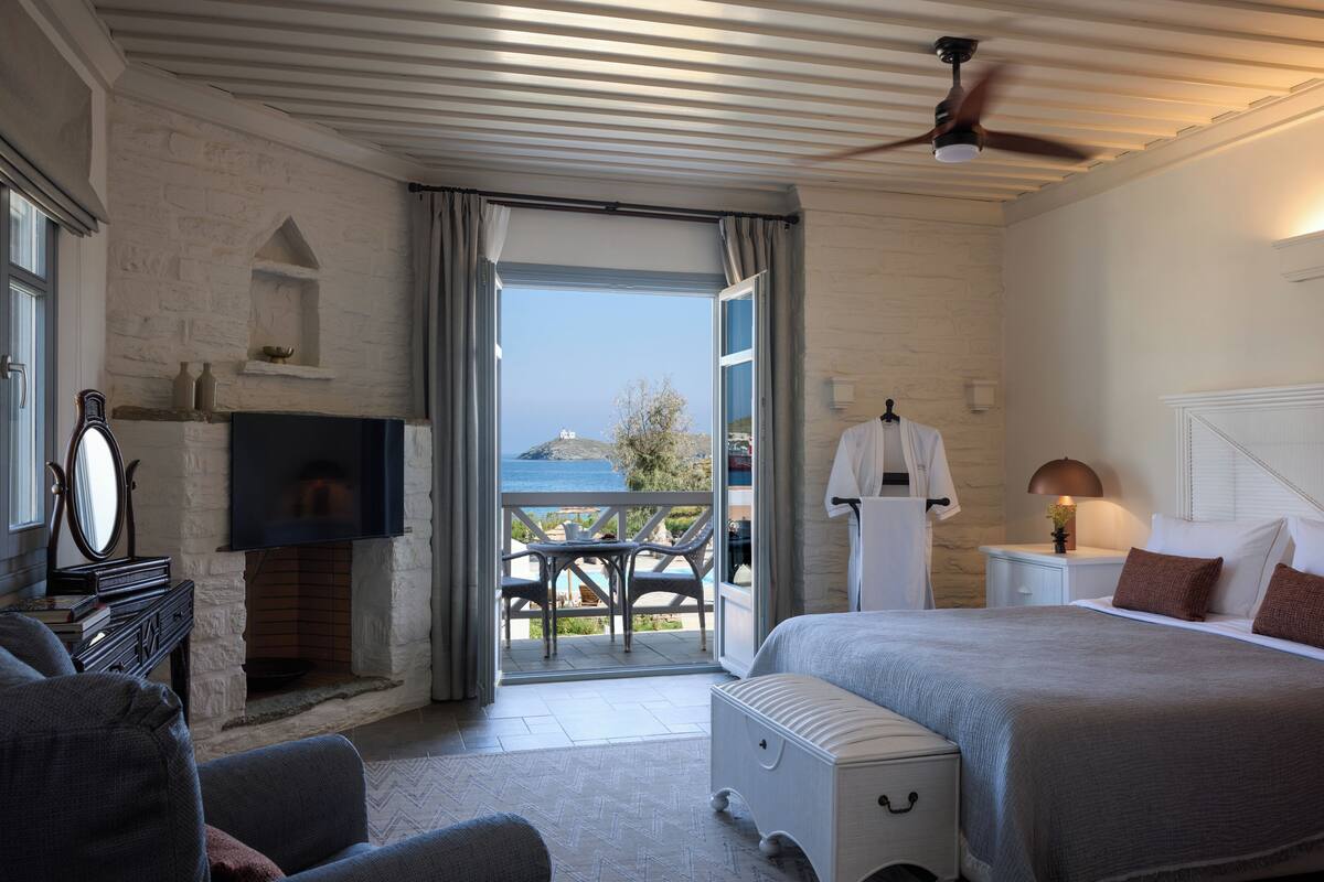 Un dormitorio con cama, televisión, chimenea, balcón con mobiliario exterior y ventilador de techo.