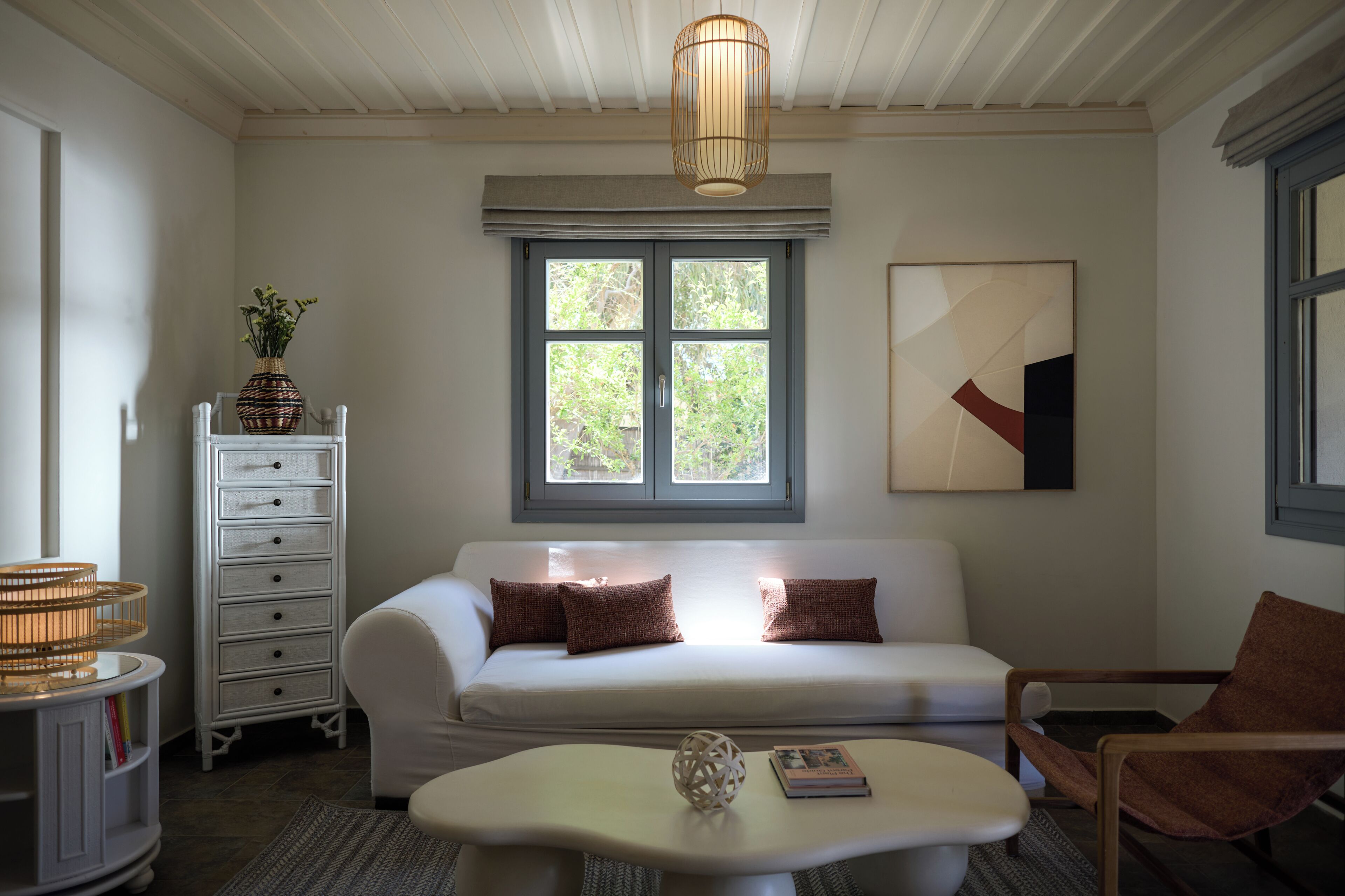 A room with a white sofa, a wooden chair, a white coffee table, a white dresser, and a window with a view of greenery.